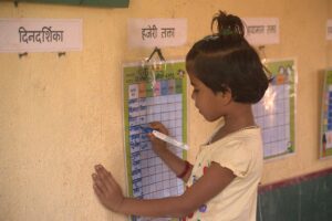 एक लहान मुलगी भिंतीवर लावलेल्या हजेरी तक्ता मध्ये नोंदी करत आहे. ती खूप लक्षपूर्वक आणि उत्साहाने तिचे नाव किंवा हजेरी दाखवत आहे. ही प्रतिमा शाळेतील नियमित हजेरी नोंदीच्या महत्वाकडे लक्ष वेधते आणि बालकांच्या शैक्षणिक सहभागाचे प्रतीक आहे.
