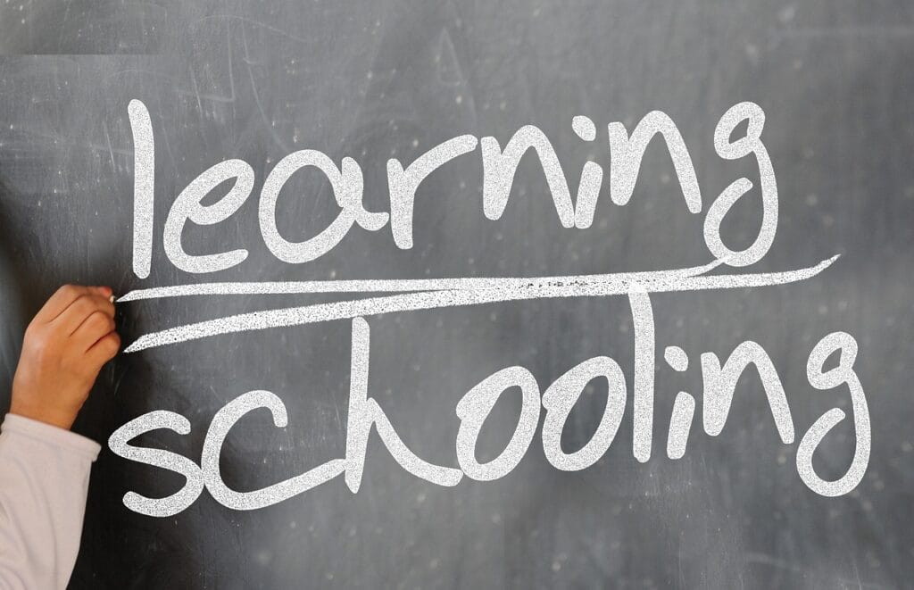 A child's hand writing the words "learning" and "schooling" on a chalkboard in white chalk.